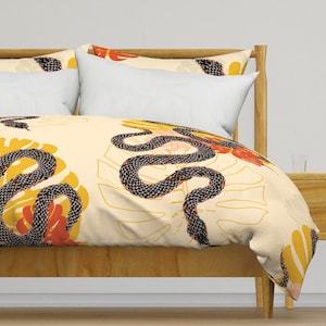Op de afbeelding: Een crèmekleurige dekbedovertrek met een patroon van een zwarte slang en gele en oranje tropische bladeren. Het dekbedovertrek ligt op een bed met een houten hoofdeinde.