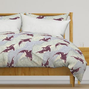 May include: A light blue and white duvet cover with a pattern of purple orcas swimming in waves. The duvet cover is on a wooden bed frame with a white pillow on top.