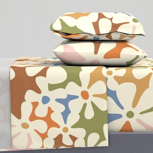 Puede incluir: Un juego de cama con fondo crema y un gran patrón floral abstracto en tonos marrones, naranjas, verdes y azules. El juego incluye una sábana bajera, una sábana encimera y dos fundas de almohada.