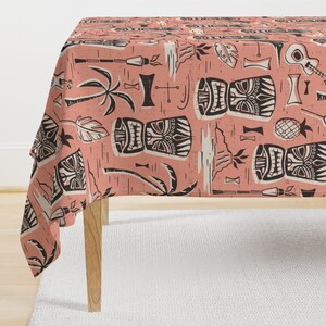 Op de afbeelding: Een koraalkleurig tafelkleed met een herhalend patroon van zwart-witte tiki-thema illustraties. Het ontwerp bevat tiki-maskers, palmbomen, gitaren en andere tropische elementen. Het tafelkleed is over een houten tafel gedrapeerd.