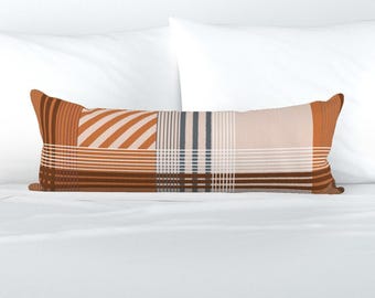 May include: A rectangular decorative pillow with a patchwork design. The pillow features various geometric patterns in shades of brown, orange, and beige. Stripes, lines, and blocks create a textured appearance. The pillow is placed on a white bed.