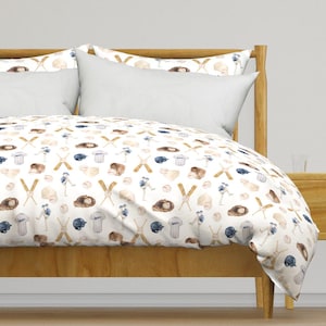 Puede incluir: Una cama con estructura de madera clara y almohadas blancas. La ropa de cama presenta un patrón repetido de bates de béisbol, guantes, pelotas de béisbol, cascos y jugadores en tonos marrón, azul y beige sobre un fondo blanco.