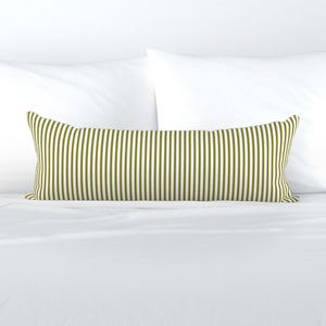 May include: Olive green and white striped lumbar pillow. The long, rectangular pillow features vertical stripes and is placed on a white bed with white pillows in the background. The pillow is designed for decorative use.