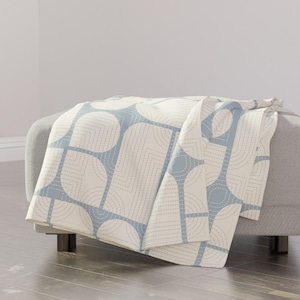 May include: A soft, white throw blanket with a geometric pattern of blue circles and lines. The blanket is folded and draped over a light gray ottoman.