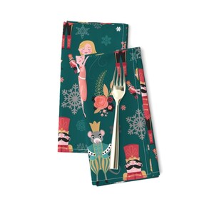 May include: A set of two teal green fabric napkins with a repeating pattern of a nutcracker, a mouse, a ballerina, snowflakes, and flowers. The napkins are folded and placed on a white surface.