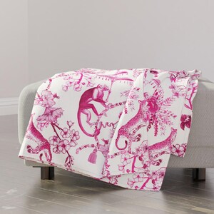 May include: A white throw blanket with a pink and white floral pattern featuring monkeys and leopards. The pattern is reminiscent of a chinoiserie design.