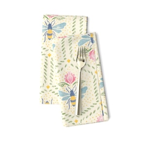 May include: A folded cream-colored cloth napkin with a bee and floral pattern. The design includes blue bees, pink flowers, yellow diamonds, and green leaf accents. A silver fork rests on the napkin.