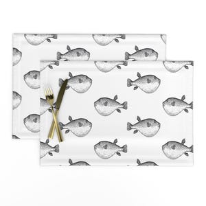 May include: White placemats with a repeating pattern of black and white pufferfish illustrations. A gold fork and knife are placed on one of the placemats. The placemats are rectangular and suitable for a dining table.