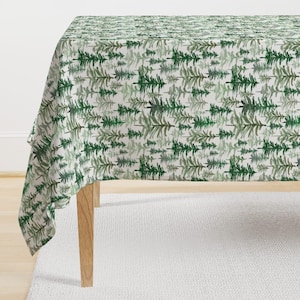 Könnte beinhalten: Eine weiße Tischdecke mit einem grünen Aquarell-Muster aus Kiefern. Die Tischdecke ist über einen Holztisch mit vier Beinen gelegt.