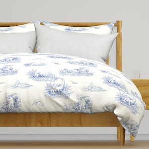 May include: A bed with a light wood frame and white bedding. The duvet cover features a blue and white toile pattern with dinosaurs, trees, and mountains. Two white pillows are visible.
