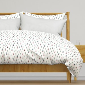 Puede incluir: Una cama con edredón y fundas de almohada blancos. El edredón presenta un patrón repetido de esquiadores y árboles verdes sobre un fondo blanco. El marco de la cama es de madera clara.