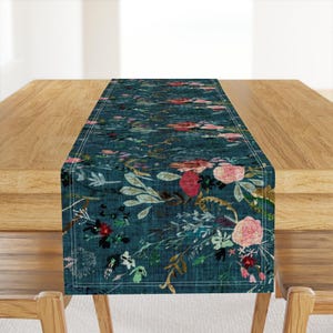 May include: A teal blue table runner with a floral pattern featuring pink, red, and white flowers. The runner has a subtle textured background and a white stitched border.