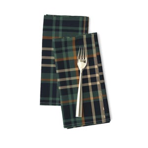 May include: Two folded cloth napkins with a plaid pattern in dark green, navy, and tan. A silver fork rests on top of one napkin. The napkins are on a white background.
