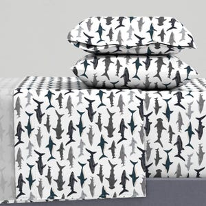 May include: Bed linen set featuring a shark pattern in grey and navy blue on a white background. The set includes a fitted sheet, a flat sheet, and two pillowcases. The shark design is repeated across the sheets and pillowcases.