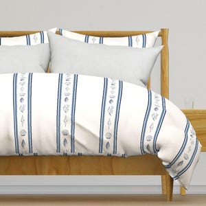 Peut inclure: Ensemble de literie à rayures blanches et bleues, orné de motifs de coquillages. Le motif présente des rayures verticales avec des accents bleus et des illustrations de coquillages. L'ensemble comprend une housse de couette et des oreillers blancs.