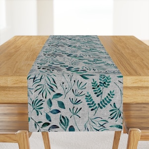 May include: A teal and gray table runner with a watercolor pattern of leaves and branches. The runner is made of linen and has a subtle texture.