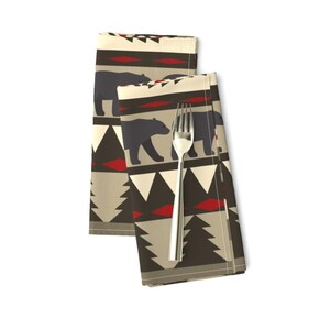 May include: Two beige fabric napkins with a repeating pattern of black bears, red and white geometric shapes, and dark green pine trees.