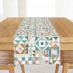May include: A white table runner with a geometric pattern in shades of brown, teal, and pink. The pattern is made up of squares, triangles, and rectangles.