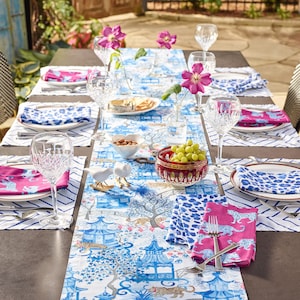 Könnte beinhalten: Ein weißer Tischläufer mit einem blauen und weißen Pagoden- und Leopardenmuster. Der Läufer liegt auf einem braunen Tisch mit weißen Platzsets und rosa Servietten. Auf dem Tisch stehen klare Weingläser und eine Schale mit Trauben.