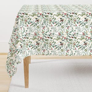 May include: A white tablecloth featuring a repeating pattern of green leaves, red berries, and gold accents. The tablecloth is draped over a wooden table, creating a festive look. The design is reminiscent of a holiday theme, suitable for a dining table.