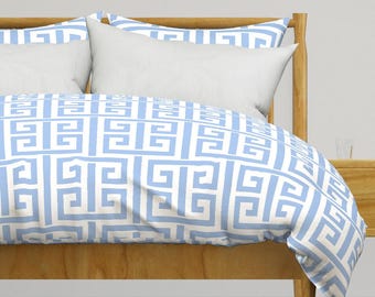 Pode incluir: Uma cama com estrutura de madeira clara, almofadas brancas e uma capa de edredão com um padrão geométrico repetido em azul claro e branco. O padrão é um desenho de chave grega. A cama está numa sala bem iluminada.