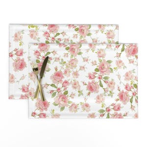 Puede incluir: Un juego de dos manteles individuales blancos con un patrón floral rosa. Los manteles individuales presentan un patrón repetido de rosas rosas y hojas verdes.