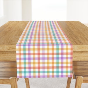May include: A colorful checkered table runner with a pastel color scheme. The runner is made of a soft, woven fabric and features a repeating pattern of orange, purple, green, and blue squares.