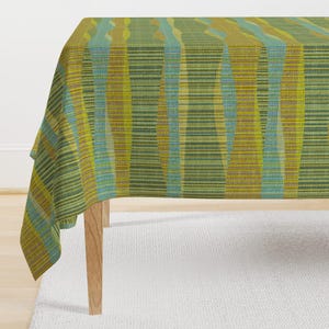 May include: A rectangular tablecloth with a geometric pattern. The design features vertical stripes in shades of green, yellow, and blue. The tablecloth is draped over a wooden table, with the corner of the cloth visible.