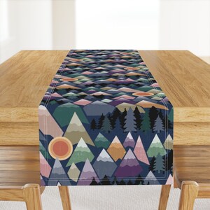 May include: A decorative table runner featuring a geometric mountain landscape design. The runner showcases a repeating pattern of colorful mountains in shades of blue, green, pink, and orange, with a dark blue background. The runner is displayed on a wooden table.