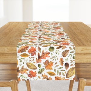 May include: A white table runner featuring a watercolor pattern of autumn leaves in shades of orange, yellow, and green. The leaves are scattered across the runner, creating a seasonal, decorative accent for a dining table or other surface.