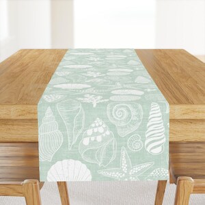 May include: A light blue table runner with a white seashell pattern. The runner features a variety of seashells, including clams, snails, and starfish.