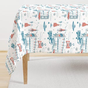 May include: A white tablecloth with a repeating pattern of retro motels, cars, flying saucers, and figures in red and blue. The tablecloth is draped over a wooden table, with the pattern visible on the surface.