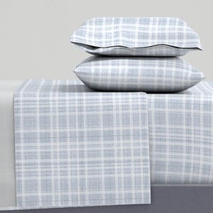 Pode incluir: Um conjunto de roupa de cama com um padrão xadrez azul claro e branco. O conjunto inclui duas almofadas empilhadas sobre um lençol dobrado, com o padrão visível nas almofadas e no lençol. Os lençóis e fronhas têm um aspeto macio e texturizado.