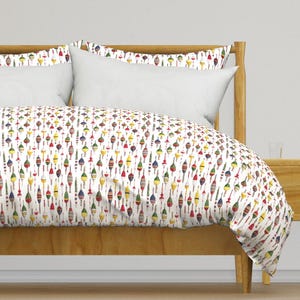 Op de afbeelding: Wit dekbedovertrek met een herhalend patroon van kleurrijke dobberstoppers. Het dekbedovertrek ligt op een houten bedframe met een wit hoofdeinde en twee witte kussens.