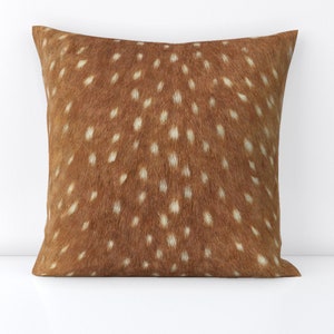 May include: Square decorative pillow with a brown and white spotted pattern, resembling deer fur. The pillow has a soft, textured appearance and is likely made of fabric. The design is evenly distributed across the surface.