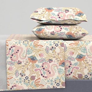 May include: A cream-colored bedding set featuring a repeating pattern of pink snakes, flowers, and foliage in shades of blue, peach, and gold. Two pillows are stacked on top of a matching bedspread, creating a cohesive and decorative look.