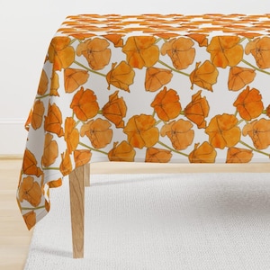 Puede incluir: Un mantel blanco con un patrón repetitivo de flores de amapola de California de color naranja. El mantel está drapeado sobre una mesa de madera.