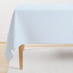 May include: A light blue and white gingham tablecloth on a wooden table. The tablecloth features a traditional checkered design, offering a simple and elegant look. The table is placed on a white rug.