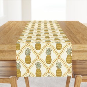 May include: A yellow and white table runner with a pineapple pattern. The pineapples are in a repeating pattern on a white background with a yellow geometric design.