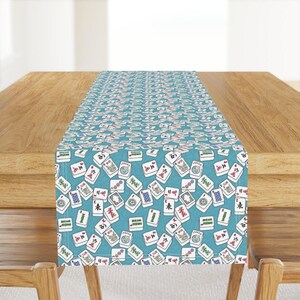 May include: A rectangular table runner featuring a repeating pattern of white mahjong tiles with various colorful symbols, characters, and designs scattered across a solid teal blue background, creating a dense and uniform graphic print.