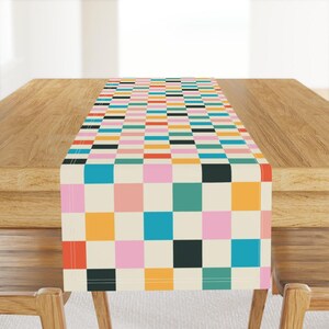 May include: A colorful checkered table runner with a cream background. The squares are in shades of pink, blue, green, orange, yellow, and black.