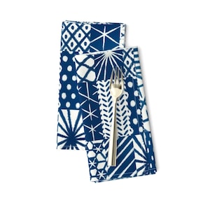 May include: Two folded blue and white patterned cloth napkins with a silver fork. The napkins feature a patchwork design with various geometric patterns. The fork is placed on top of the napkins.