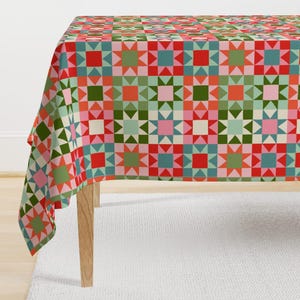 Könnte beinhalten: Eine bunte Tischdecke mit geometrischem Muster. Das Design zeigt Quadrate, Dreiecke und Sternformen in Rot-, Grün-, Rosa- und Blautönen. Die Tischdecke liegt auf einem Holztisch.