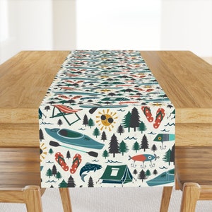 May include: A white table runner with a colorful pattern of camping and fishing themed icons. The pattern includes trees, a sun, a kayak, a tent, fish, flip flops, and fishing lures.