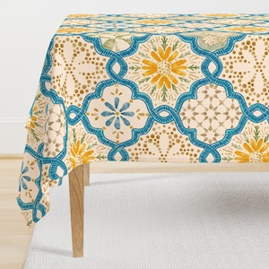 May include: A rectangular tablecloth with a repeating pattern of blue, yellow, and beige floral and geometric designs. The tablecloth is draped over a wooden table, showcasing the intricate pattern and color scheme. The design includes stylized flowers and interconnected shapes.