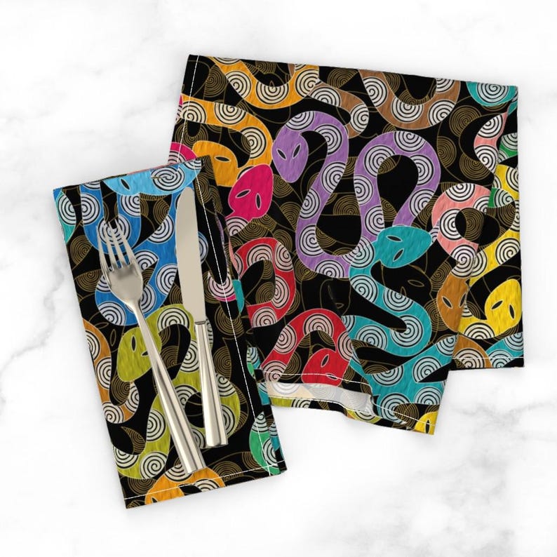 May include: Two black fabric napkins with a colorful snake pattern. The snakes are outlined in gold and have a swirling pattern. The napkins are folded and placed on a white surface.