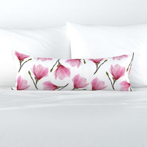 May include: A long, rectangular pillow with a white background and a repeating pattern of pink magnolia flowers. The flowers have green stems and buds. The pillow is placed on a white bed with two white pillows.
