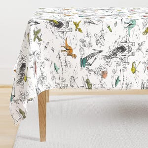 May include: White tablecloth with a whimsical black and white illustration of dinosaurs, planes, and other objects. The design includes pops of color with green, orange, and yellow accents. The tablecloth is draped over a wooden table.