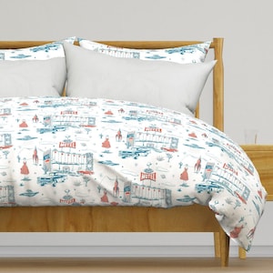 May include: White duvet cover and pillowcases with a retro motel print. The design features illustrations of motels, cars, rockets, and figures in red and blue. The bedding is set on a wooden bed frame.