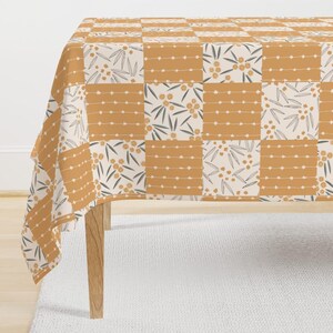 May include: A mustard yellow and cream-colored tablecloth with a patchwork design. The squares feature alternating patterns of dotted lines, floral sprigs, and leaf motifs. The tablecloth is draped over a wooden table.